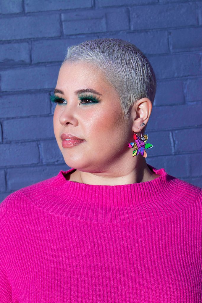 April Watson wearing green eyelashes with a pink knit jumper, brightly coloured earrings and a shaved head.