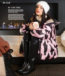 A brunette model sitting beside a fireplace holding a cup, wearing a white beanie and pink and black puffy jacket.