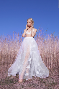 A blonde model wearing a white bralette with a long white flowy skirt in front of long grass.